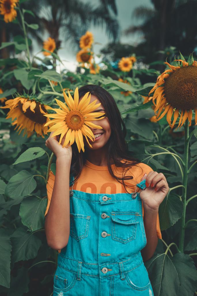 Woman of color wearing blue overalls holding up a sunflower and smiling in a field of sunflowers